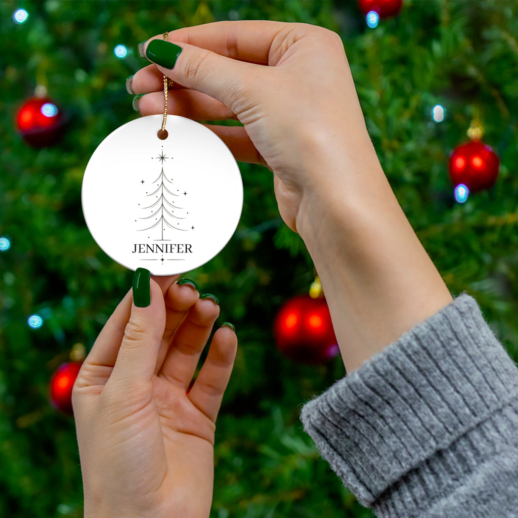 Hand holding a round personalized Christmas tree ornament in front of a Christmas tree
